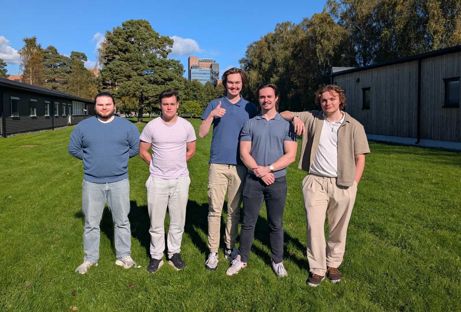 Studentene bak prosjektet står samlet på en gressplen utenfor Universitetet i Agder i Kristiansand på en solrik dag.